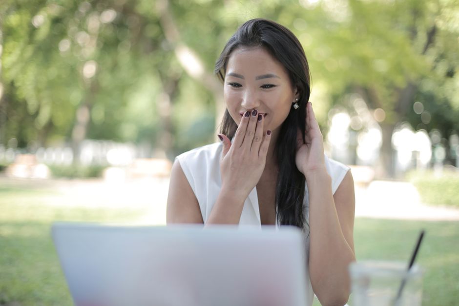 An Asian woman reacts with surprise while using a laptop in a sunny park.