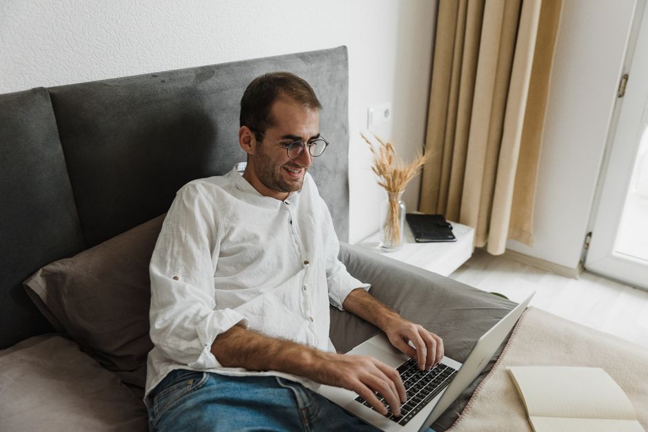 A man wearing eyeglasses working on his laptop while laying in bed with a smile.