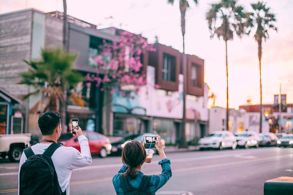 Back view anonymous couple in casual clothes taking photos on mobile phones while standing on street in tropical modern city at twilight