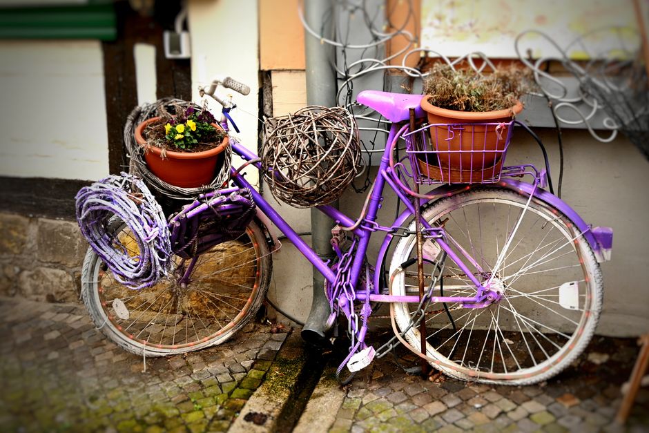 A purple vintage bicycle adorned with flower pots and artistic wicker decorations, leaning against a building.