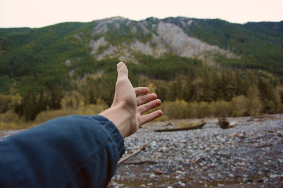 A person's hand reaching out towards a scenic mountain landscape covered with greenery and rocks.