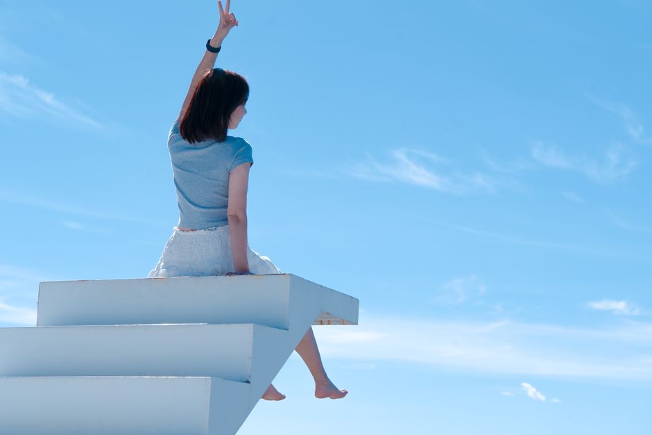 A woman sits on white stairs with blue sky background, reaching upward, embracing freedom.