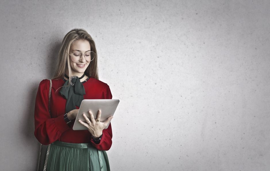 A beautiful woman in glasses standing against a gray wall, engaging with a tablet.