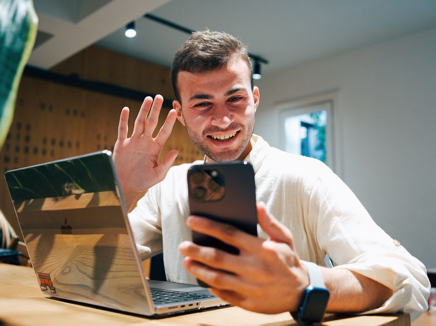 Smiling young man making a video call on his smartphone while sitting with a laptop in a modern office.