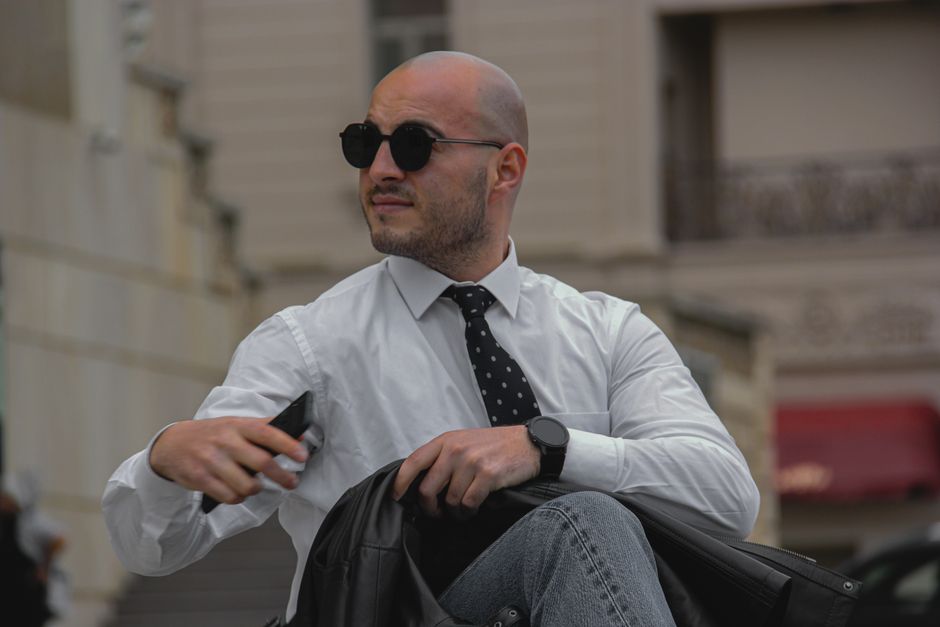 Businessman in sunglasses and tie sitting outdoors, exuding confidence.