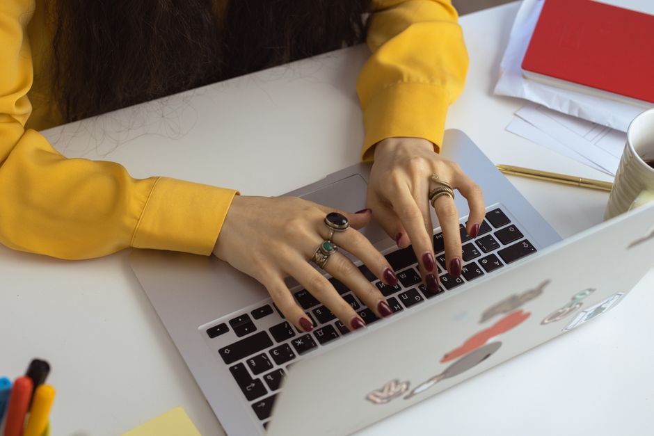 Close-up of a woman typing on a laptop, showcasing red nails and rings, wearing a yellow long sleeve indoors.