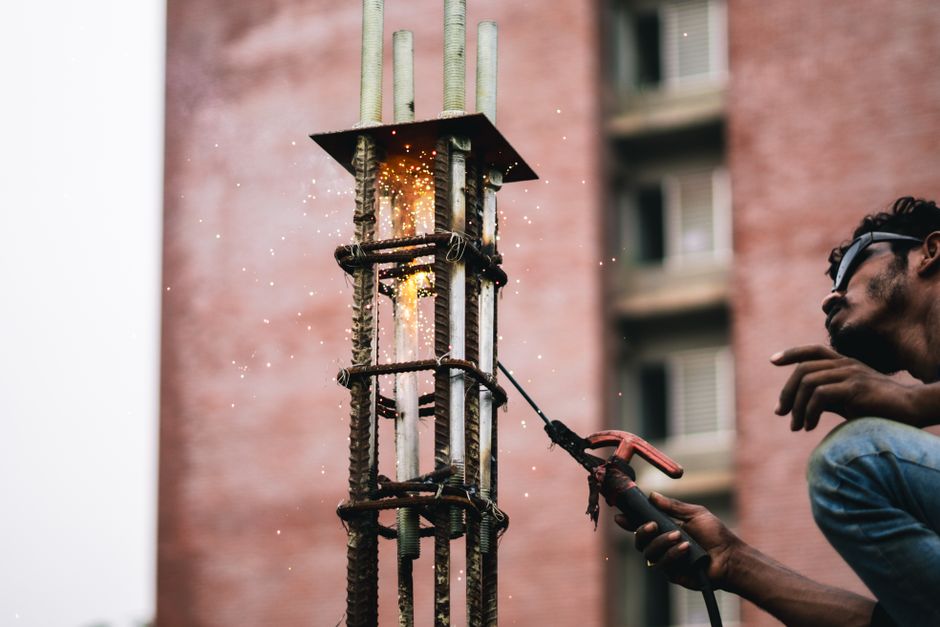 A construction worker welds iron rods with sparks flying against a brick wall backdrop.