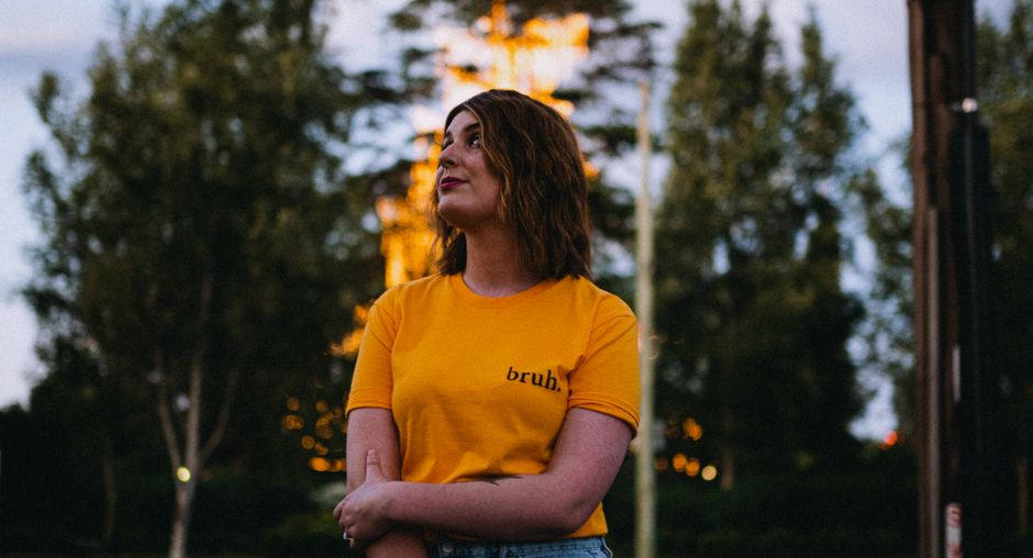 A woman in a yellow shirt stands outdoors at twilight, surrounded by trees and soft lighting.