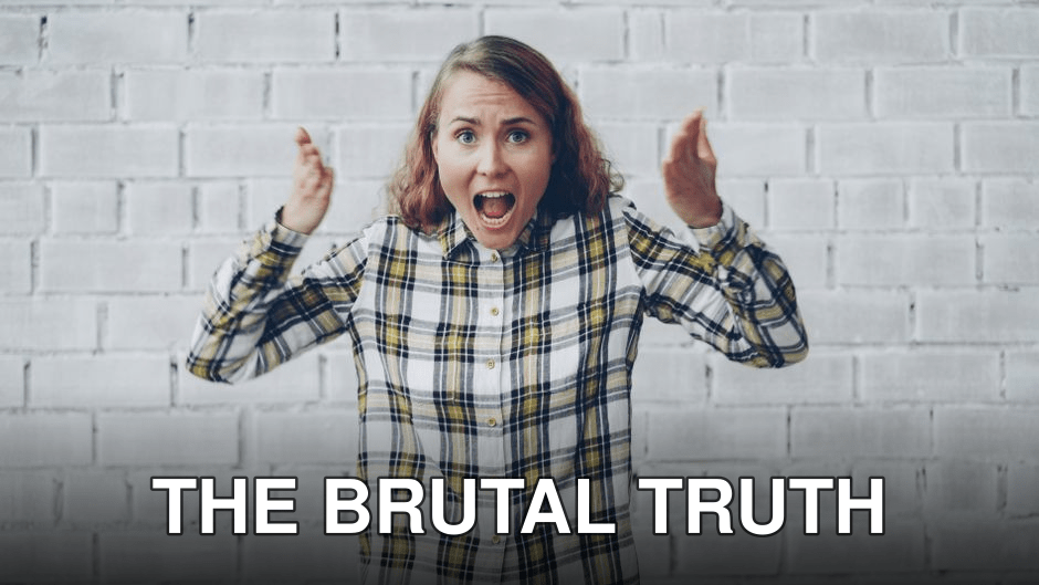 Shocked woman with raised hands in plaid shirt, standing indoors against a white brick wall.
