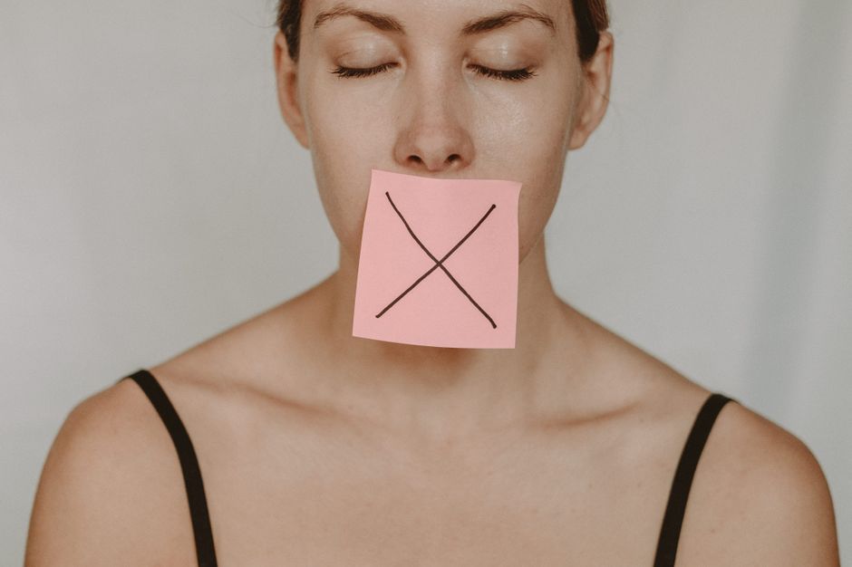 Young woman in black lingerie with closed eyes showing pink sticker with black cross against violence and abuse on lips on white background in light studio