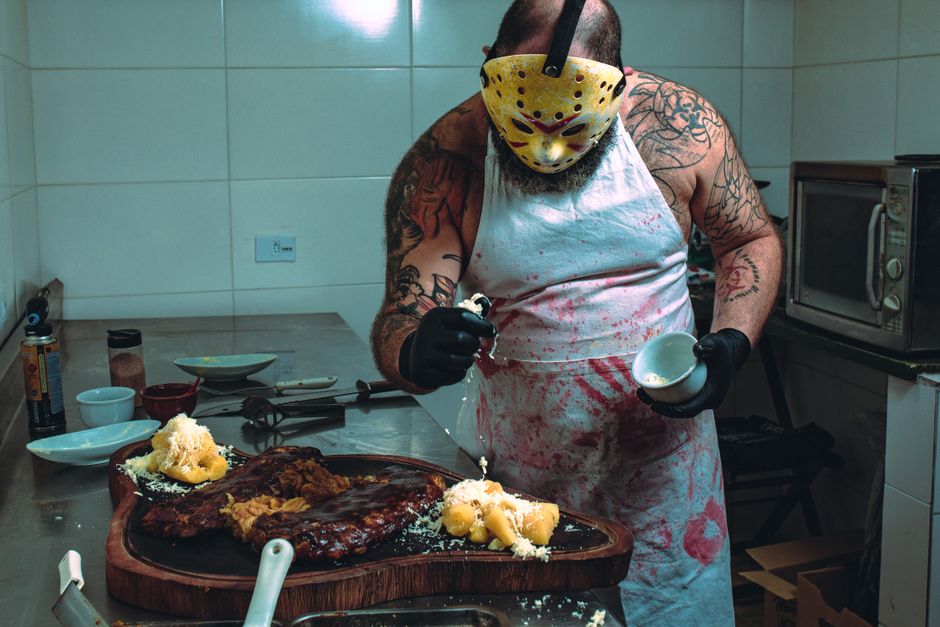 A tattooed man in a horror mask garnishes food in a kitchen. Creepy culinary art.
