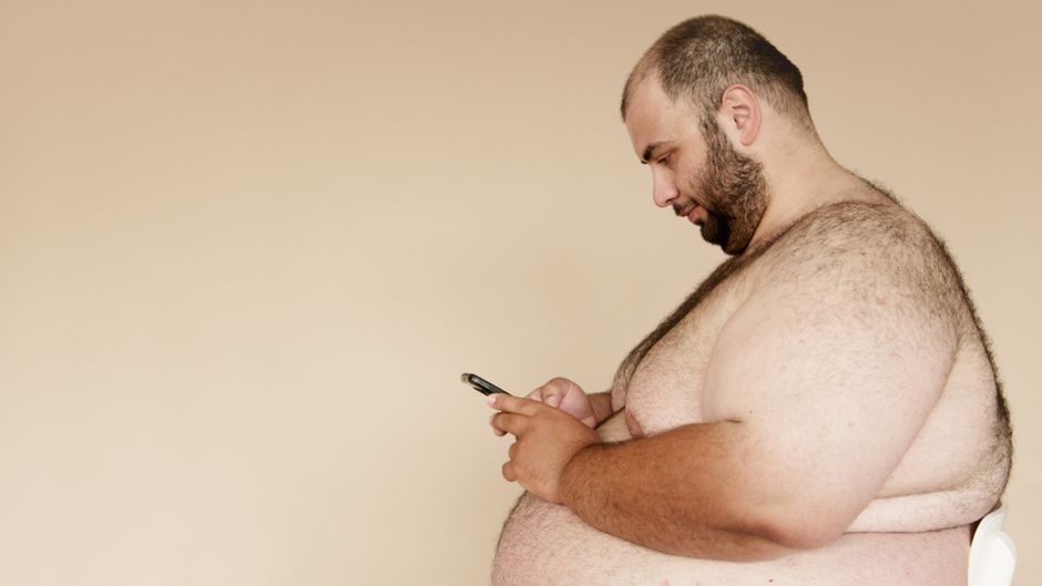Side view of an overweight man using a smartphone indoors, showcasing modern technology usage.
