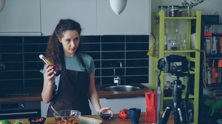 A woman filming a cooking vlog in her kitchen holding a condiment bottle.