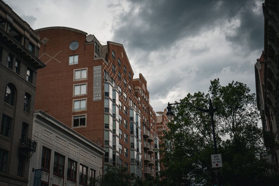 View of an urban cityscape under a dramatic cloudy sky, featuring tall buildings and street elements.