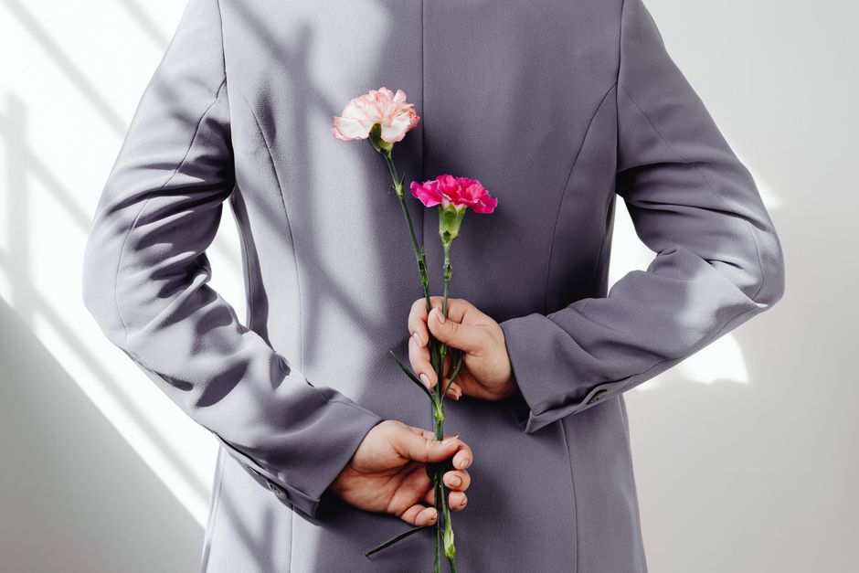 Person wearing gray suit holding two vibrant carnations behind their back.