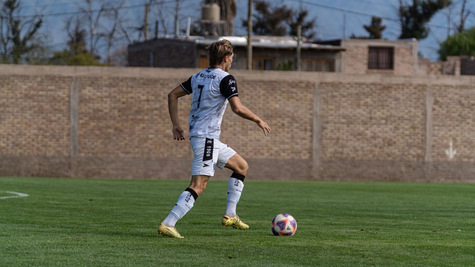 Soccer player dribbles the ball on a grassy field against a residential backdrop. Action captured during the day.