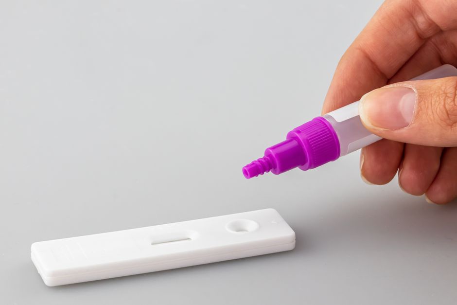 Close-up of a hand applying liquid on a rapid test kit, against a white surface.
