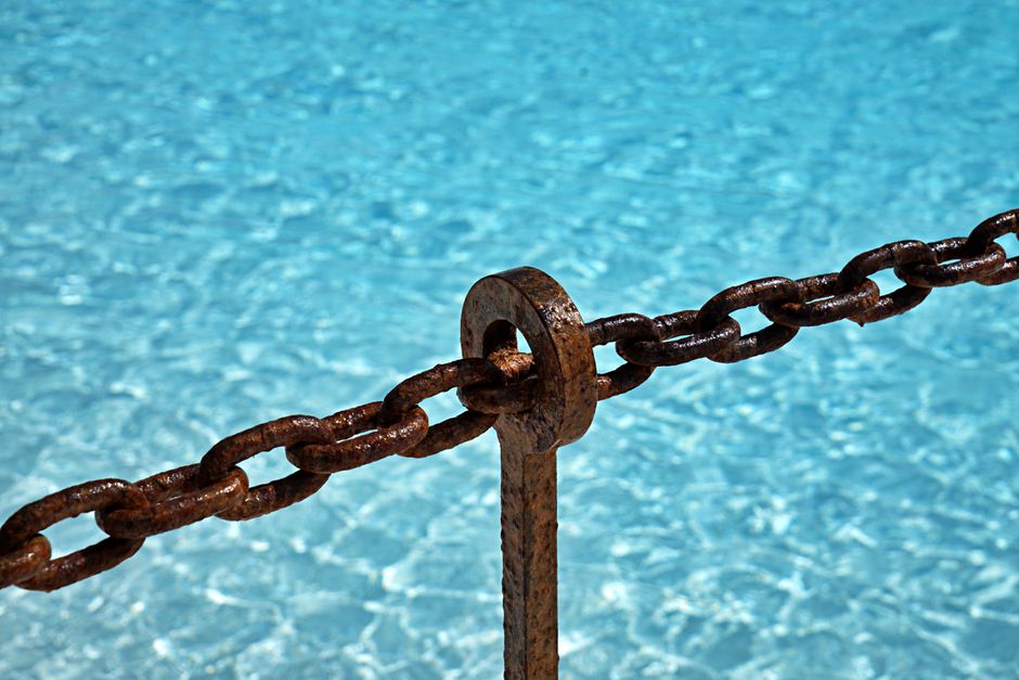 Close-up of a rusty chain suspended above sparkling blue water, capturing a contrast between antique metal and clear liquid.