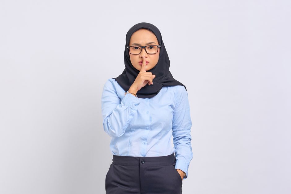 Asian Muslim woman in hijab making a quiet gesture with a serious expression, wearing eyeglasses against a white background.
